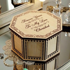 Könnte beinhalten: Eine achteckige Schatulle aus hellem Holz mit einem dunkelbraunen, lasergravierten Rand aus Herzen und Schnörkeln. Der Deckel trägt den Schriftzug "Forever Yours To My Love Cherished". Die Seiten haben quadratische Paneele mit Herzmuster.