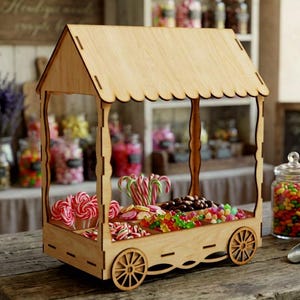 Puede incluir: Un expositor de dulces de madera clara con forma de carrito, techo festoneado y cuatro ruedas. El carrito cuenta con varios compartimentos llenos de diversos dulces coloridos, incluyendo piruletas rojas y blancas, ositos de goma y chocolate.