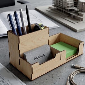 May include: A light-colored wooden desk organizer with a tall compartment holding three pens and a small measuring tape, a middle section containing a gray business card with the text Archicorp, and a lower section holding a stack of green sticky notes.