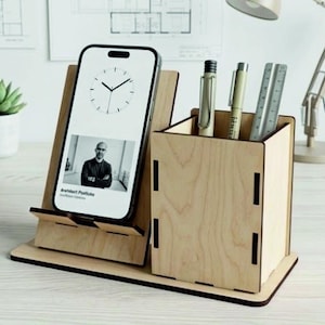 Op de afbeelding: Een bureau-organizer van lichtgekleurd hout met een schuine smartphonehouder aan de linkerkant en een vierkante pennenhouder aan de rechterkant. De telefoon toont een klok en een zwart-wit portret met de tekst "Arendbert Portfolio".