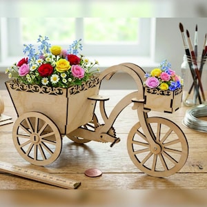 May include: A light-colored wooden tricycle planter featuring a large rear basket and a smaller front basket, both filled with colorful artificial flowers including red, yellow, pink, and blue blossoms. A wooden ruler is visible in the foreground.