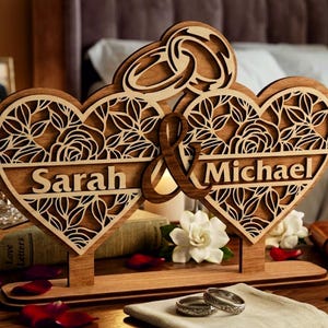 Op de afbeelding: Een lasergesneden decoratief houten bord met twee verbonden harten met ingewikkelde bloemmotieven. De namen Sarah en Michael zijn gegraveerd op horizontale balken over de harten, verbonden door een ampersand. Bovenaan bevinden zich twee ringen.