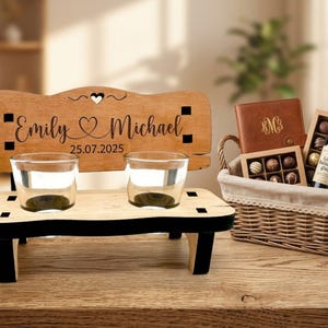 May include: A wooden bench-shaped holder with two glass cups, engraved with the names Emily and Michael and the date 25.07.2025. Beside it sits a wicker basket containing a brown leather-bound notebook, a box of chocolates, and a bottle of glaze.