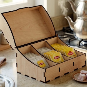 May include: A light-colored wooden tea box with a hinged lid and three internal compartments. The box contains multiple Lipton tea bags in yellow and white paper packaging. The edges of the wooden panels feature dark, laser-cut detailing.