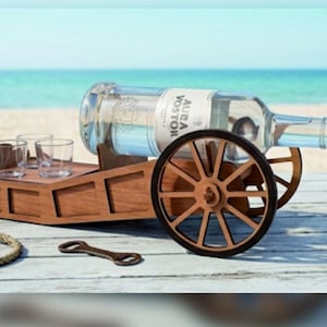 May include: A wooden cannon-shaped bottle holder with two large spoked wheels, designed to cradle a clear glass bottle labeled AURA VOSTOR. The structure includes a flat platform with two small clear glass shot glasses and a bottle opener.