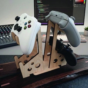 May include: A light-colored wooden stand designed to hold three video game controllers. The stand features cut-out circular details and supports a white controller on the left, with a grey and a black controller positioned on the right side.