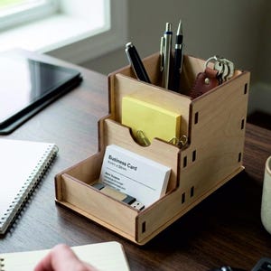 May include: A tiered wooden desk organiser with a light brown finish. The top section holds pens and a keychain, the middle section contains yellow sticky notes, and the bottom section holds business cards with the text 91mm x 55mm and a USB drive.