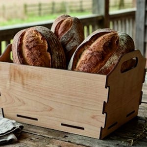 Puede incluir: Una caja rectangular de madera clara con juntas entrelazadas y asas recortadas, que contiene tres hogazas de pan artesanal redondas y crujientes con superficies doradas y marcas de corte visibles en la parte superior.