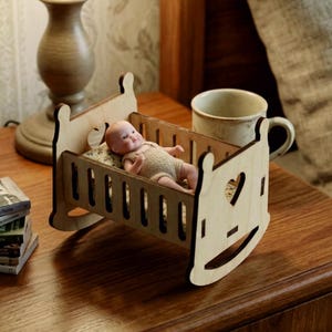 May include: A small wooden rocking cradle featuring heart-shaped cutouts on the headboard and footboard. Inside the cradle lies a miniature doll wearing a beige knitted outfit, resting on a small patterned pillow and mattress set.