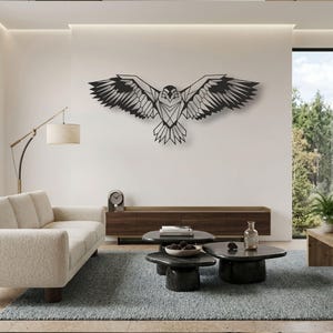 May include: A black metal wall art piece featuring a geometric eagle with wings fully spread in flight. The design consists of sharp, angular lines and shapes that form the silhouette of the bird, mounted centrally on a plain white wall.