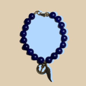 May include: A dark blue beaded bracelet with a gold clasp and charm. The charm features an anchor and a feather. The bracelet is made of round, polished beads. The background is a light blue and beige.
