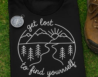 Puede incluir: Camiseta negra con un gráfico blanco de montañas, árboles, un río y el sol, con el texto "get lost to find yourself". También se ven una brújula plateada y una bota de senderismo marrón.