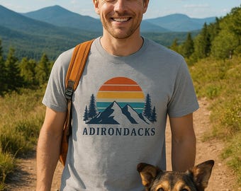 Puede incluir: Un hombre y un perro en un sendero. El hombre lleva una camiseta gris con un gráfico de montaña y puesta de sol y la palabra "ADIRONDACKS". El perro es marrón y negro con un collar rojo. Montañas y árboles en el fondo.