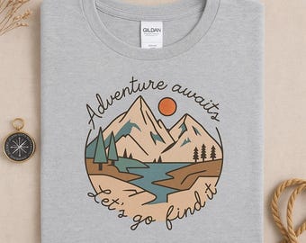 May include: A light gray t-shirt featuring a circular graphic with a mountain range, lake, and trees. The design includes the text "Adventure awaits, Let's go find it" in a handwritten style. The shirt is folded.