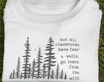May include: White t-shirt with a crew neck, featuring a forest graphic and the text "not all classrooms have femr walls. go learn from the wild." The design is in dark gray. The shirt is folded on a green surface.