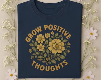 May include: A navy blue t-shirt with a floral design and the words "GROW POSITIVE THOUGHTS" in gold. The floral design features yellow flowers and green leaves. The shirt is surrounded by white flowers and greenery.