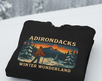 Puede incluir: Sudadera con capucha negra con un gráfico de un excursionista en un paisaje invernal y el texto "ADIRONDACKS WINTER WONDERLAND". El diseño incluye una puesta de sol, montañas y pinos.