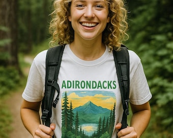 May include: A white t-shirt with the word "ADIRONDACKS" in green and black, featuring a mountain and lake scene. The person is wearing a black backpack and smiling in a forest setting.