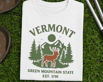 May include: White t-shirt with a green and brown graphic featuring the word "VERMONT" above a mountain scene with trees, a deer, and the text "GREEN MOUNTAIN STATE EST- 1791". The shirt is folded on green grass.