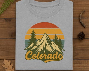 Puede incluir: Camiseta gris claro con un diseño retro de montañas, pinos y una puesta de sol en tonos naranja, amarillo y verde azulado. La palabra "Colorado" está escrita en una fuente estilizada. La camiseta está doblada sobre una superficie de madera.