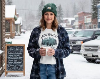 Puede incluir: Una persona lleva una camiseta blanca con el texto "ADIRONDACKS" sobre un gráfico de un oso y montañas. También lleva un gorro verde, vaqueros azules y una camisa a cuadros azul. El fondo es una escena de calle nevada.
