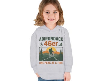 Puede incluir: Una sudadera con capucha gris claro con el texto "ADIRONDACK 46er" y un gráfico de un excursionista en una montaña con una puesta de sol. La frase "ONE PEAK AT A TIME" está debajo. La sudadera con capucha la lleva un niño pequeño.