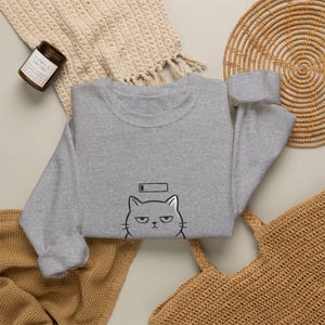 Puede incluir: Una sudadera gris jaspeado de cuello redondo con una ilustración de líneas negras de una cara de gato gruñón y un icono de batería baja situado justo encima de la cabeza del gato. La sudadera tiene mangas largas y puños acanalados.