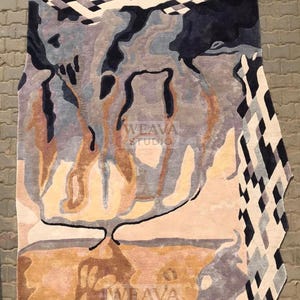 May include: Abstract-shaped rug with a unique design. The rug features a blend of colors, including shades of brown, blue, gray, and cream. The design incorporates organic shapes and geometric patterns. The text "WEAVA STUDIO" is visible on the rug.