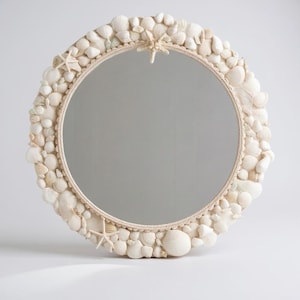 Puede incluir: Espejo redondo con un marco decorado con diversas conchas marinas y estrellas de mar en tonos blancos y crema. Un borde similar a una cuerda bordea la superficie reflectante, creando un tema costero o náutico.