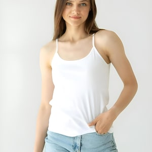 Könnte beinhalten: Ein weißes Camisole-Top mit dünnen Spaghettiträgern. Das Top wird mit hellblauen Jeans getragen. Das Model steht vor einem weißen Hintergrund.