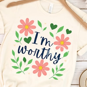 May include: Cream-colored t-shirt with the text "I'm worthy" in navy blue script, surrounded by pink flowers, green leaves, and hearts. The shirt is hanging on a wooden hanger.