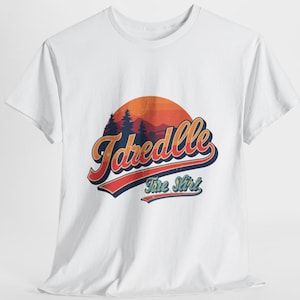 Può includere: T-shirt bianca con un design retrò. Il design presenta la parola "Jaredlle" in corsivo arancione e blu, sopra le parole "The Shirt". Sono inclusi anche un tramonto e una grafica di montagna.