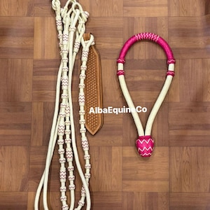 Puede incluir: Un juego de brida y riendas de caballo color crema con detalles en rosa. Las riendas presentan costuras y cuentas rosas decorativas. La brida tiene un diseño tejido rosa y blanco. El conjunto incluye una pieza de cuero con un patrón tejido.