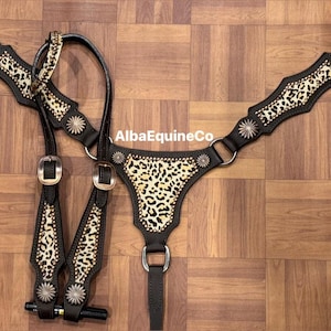 Könnte beinhalten: Schwarzes und hellbraunes Pferdegeschirr mit Leopardenmuster. Das Geschirr hat silberfarbene Akzente und goldfarbene Schnallen. Das Design umfasst einen Brustriemen, einen Kopfriemen und Zügel. Das Produkt wird auf einer Holzoberfläche präsentiert.