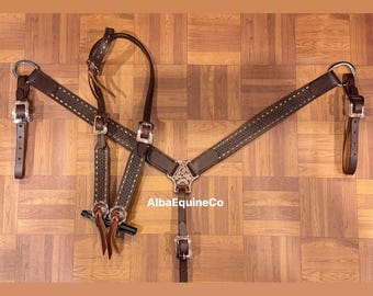 Puede incluir: Un petral de cuero marrón para caballos con herrajes plateados y costuras decorativas. El petral tiene una pieza central en forma de estrella y está diseñado para distribuir la presión de manera uniforme. El texto "AlbaEquineCo" es visible.