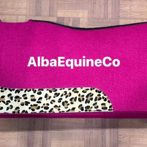 Peut inclure: Un tapis de selle magenta avec une bordure noire et des motifs léopard. Le texte "AlbaEquineCo" est imprimé en blanc. Le tapis est rectangulaire avec des coins arrondis.