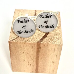 May include: A pair of silver cufflinks with white enamel and black text that reads "Father of the Bride".
