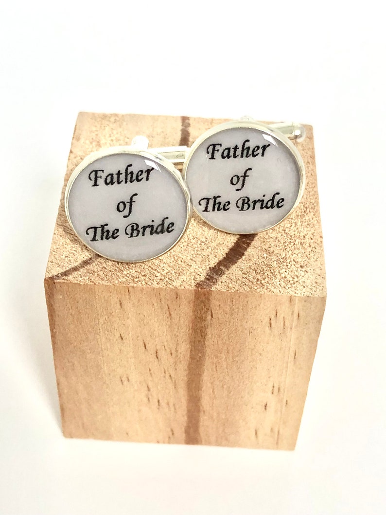 May include: A pair of silver cufflinks with white enamel and black text that reads "Father of the Bride".