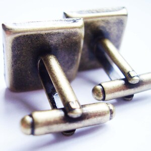 May include: A pair of gold-toned cufflinks with a square design. The cufflinks have a simple, elegant style.