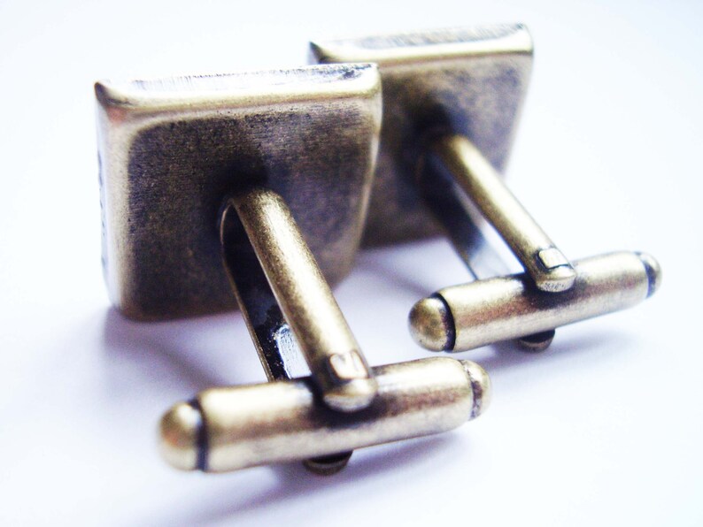 May include: A pair of gold-toned cufflinks with a square design. The cufflinks have a simple, elegant style.