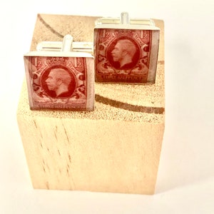 Könnte beinhalten: Ein Paar silberne Manschettenknöpfe mit roten Briefmarken, die ein Porträt von König Georg V. zeigen. Die Briefmarken sind in klarem Harz eingebettet.