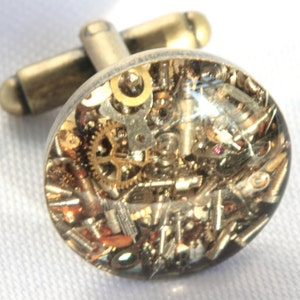 Op de afbeelding: Gouden manchetknopen met een transparante harskoepel die een verscheidenheid aan kleine tandwielen en metalen onderdelen bevat.