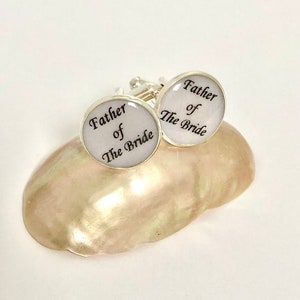 May include: A pair of silver cufflinks with white enamel and black lettering. The cufflinks say "Father of the Bride".