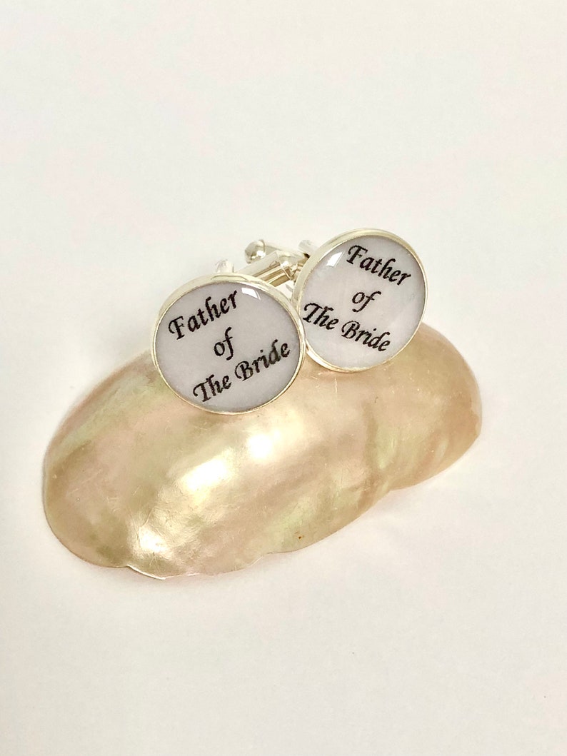 May include: A pair of silver cufflinks with white enamel and black lettering. The cufflinks say "Father of the Bride".