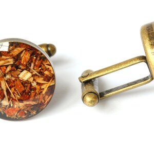 May include: A pair of cufflinks with a round, brown, and gold design. The cufflinks feature a circular, clear resin inlay with wood shavings embedded inside.