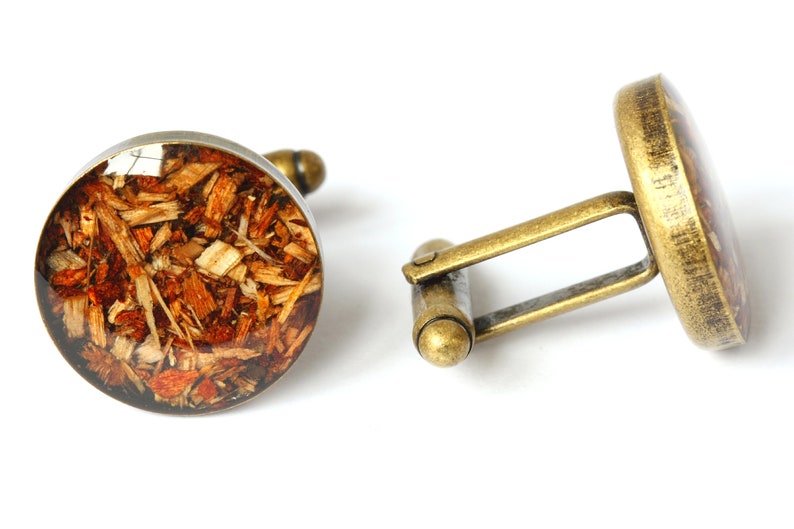 May include: A pair of cufflinks with a round, brown, and gold design. The cufflinks feature a circular, clear resin inlay with wood shavings embedded inside.