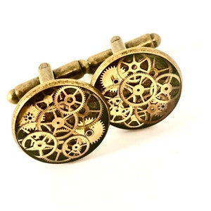 May include: A pair of gold-toned cufflinks with a circular design featuring a pattern of interlocking gears. The gears are a dark brown colour and are set against a gold background.