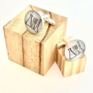 May include: A pair of silver cufflinks with a design of two beakers and a graduated cylinder. The cufflinks are displayed on two small wooden blocks.