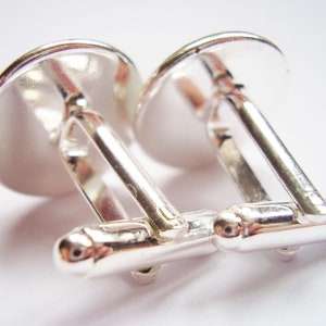 May include: A pair of silver cufflinks with a round, smooth, silver top. The cufflinks are connected by a silver bar.