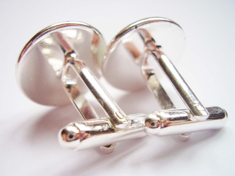 May include: A pair of silver cufflinks with a round, smooth, silver top. The cufflinks are connected by a silver bar.
